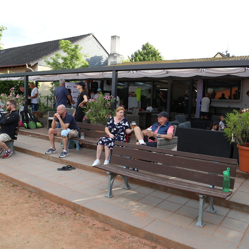 Menschen sitzen und stehen auf einer Terrasse mit Blumenbeeten und B&auml;nken, umgeben von einem einladenden Ambiente.