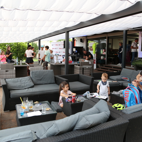 Belebte Lounge mit Sitzgruppen, Kindern beim Spielen und G&auml;sten, die Getr&auml;nke genie&szlig;en, unter einem Sonnensegel.