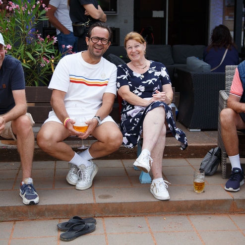 Vier Personen sitzen auf einer Bank im Freien, Getr&auml;nke in der Hand, umgeben von Blumen und Pflanzen.