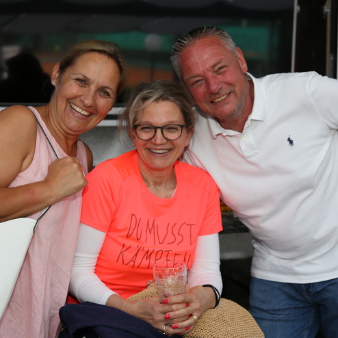 Drei Personen l&auml;cheln in einer lockeren Atmosph&auml;re, eine Frau tr&auml;gt ein leuchtend pinkes T-Shirt mit dem aufgedruckten Spruch.