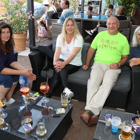 Vier Personen sitzen entspannt auf einer Terrasse mit Getr&auml;nken auf einem Tisch, umgeben von Pflanzen und Besuchern.