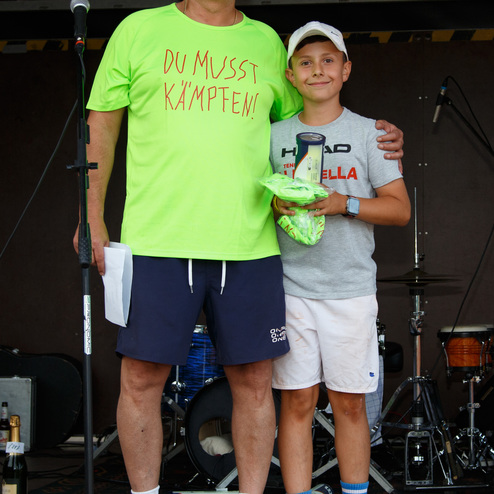 Zwei Personen stehen vor einem Mikrofon auf einer B&uuml;hne, der Erwachsenen tr&auml;gt ein leuchtend gr&uuml;nes T-Shirt mit Aufschrift.