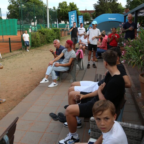 Gruppe von Menschen auf einer Terrasse bei einem Tennisplatz, einige schauen dem Spiel zu und andere unterhalten sich.