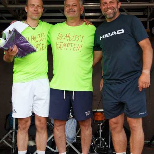 Drei M&auml;nner in Sportkleidung mit T-Shirts stehen l&auml;chelnd auf einer B&uuml;hne, hinter ihnen eine Band und Musikinstrumente.