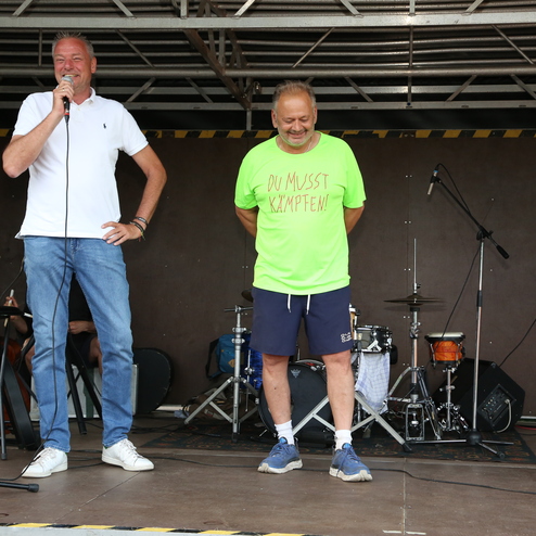 Zwei M&auml;nner stehen auf einer B&uuml;hnenplattform, ein Mann spricht ins Mikrofon, der andere tr&auml;gt ein leuchtend gr&uuml;nes Shirt.