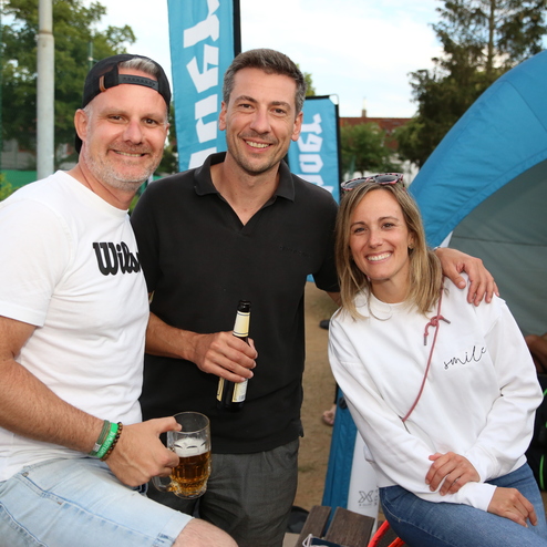 Drei Personen stehen l&auml;chelnd zusammen, einer h&auml;lt ein Getr&auml;nk, im Hintergrund ist eine Sportveranstaltung erkennbar.