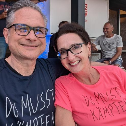 Paar sitzt in einem Caf&eacute;, beide tragen T-Shirts mit der Aufschrift &bdquo;DU MUSST K&Auml;MPFEN&ldquo;. Im Hintergrund sind weitere Personen zu sehen.