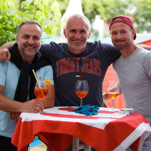 Drei M&auml;nner stehen freundlich zusammen an einem Tisch mit Getr&auml;nken in Au&szlig;enszenario. Im Hintergrund ist ein rotes H&auml;uschen sichtbar.