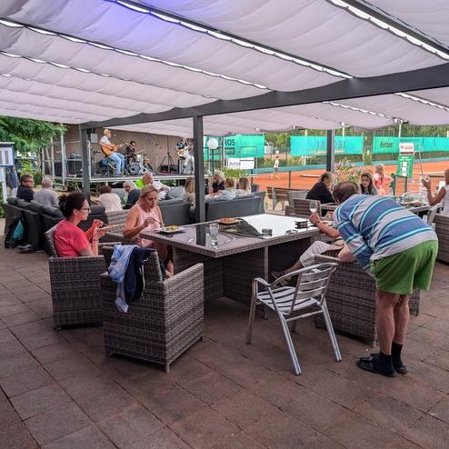 Menschen sitzen an Tischen unter einer &Uuml;berdachung und genie&szlig;en die Atmosph&auml;re eines Tennisplatzes im Hintergrund.