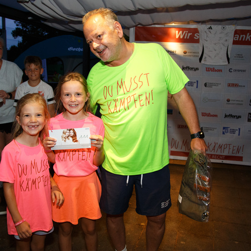 Drei Personen stehen im Vordergrund: zwei M&auml;dchen in leuchtenden T-Shirts und ein Mann mit einer Urkunde.