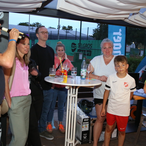 Gruppenszene mit sechs Personen an einem Stehtisch, Getr&auml;nke in der Hand, bei einem Event im Freien.