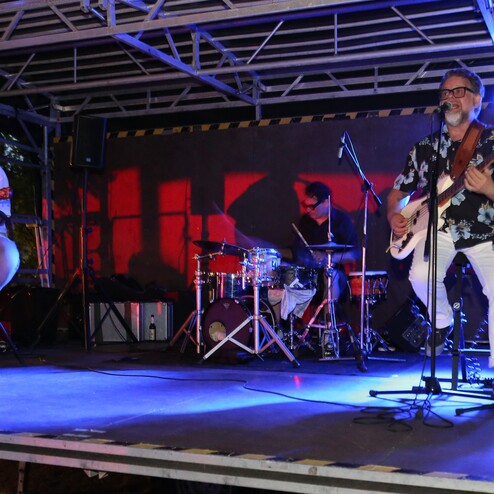 Drei Musiker spielen auf einer B&uuml;hne: ein S&auml;nger mit E-Gitarre, ein Akustikgitarrist und ein Schlagzeuger.