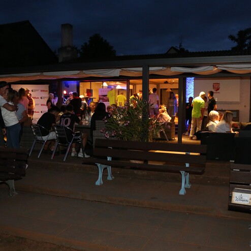 Menschen stehen und sitzen bei einer Veranstaltung im Freien vor einem beleuchteten Geb&auml;ude mit Gartenm&ouml;beln.