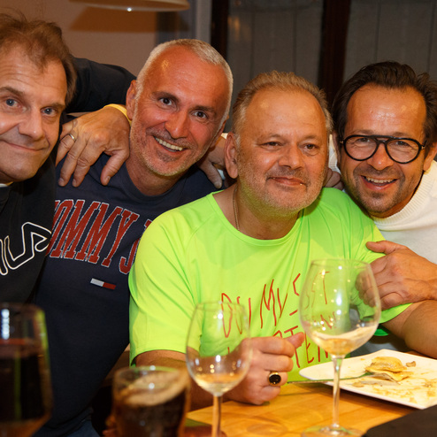 Vier M&auml;nner posieren l&auml;chelnd um einen Tisch mit Gl&auml;sern und Tellern in einem gem&uuml;tlichen Raum.