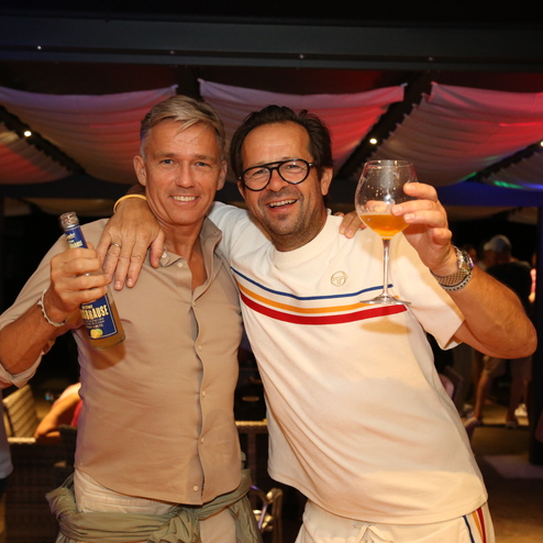 Zwei M&auml;nner posieren l&auml;chelnd mit Getr&auml;nken auf einer sommerlichen Feier unter einer festlich dekorierten &Uuml;berdachung.