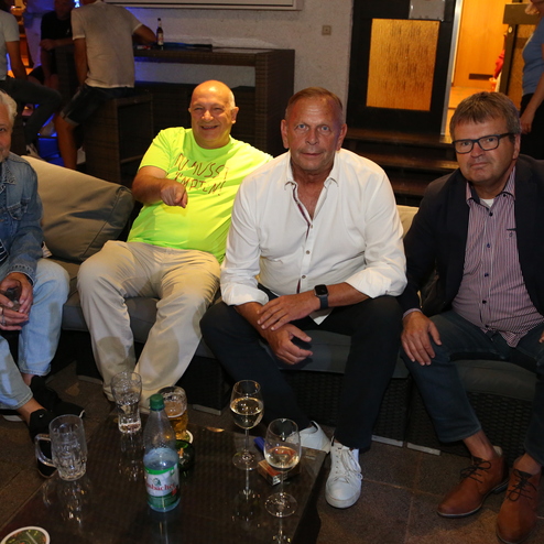 Gruppe von vier M&auml;nnern sitzt entspannt auf einer Couch in einer Bar, umgeben von Getr&auml;nken und ged&auml;mpftem Licht.