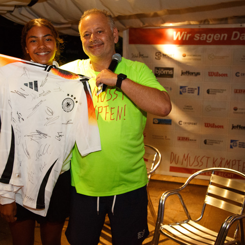 Ein junger Mensch h&auml;lt ein signiertes Fu&szlig;balltrikot, w&auml;hrend eine Person in Neon-T-Shirt daneben l&auml;chelt.