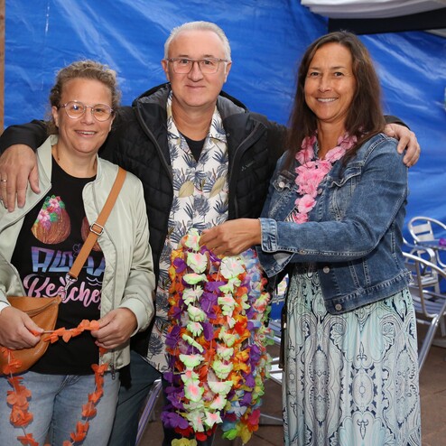 Drei Personen mit Blumenkr&auml;nzen l&auml;cheln vor einer blauen Wand, Hintergrund mit Tischen und St&uuml;hlen.