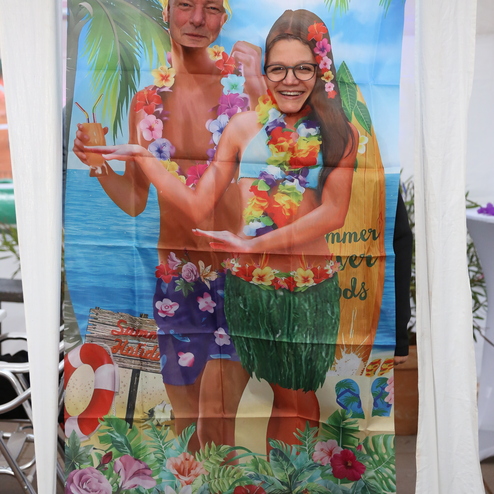 Pappaufsteller mit zwei Personen in sommerlicher Kleidung vor Strandkulisse und Palmen, umrahmt von bunten Blumen.