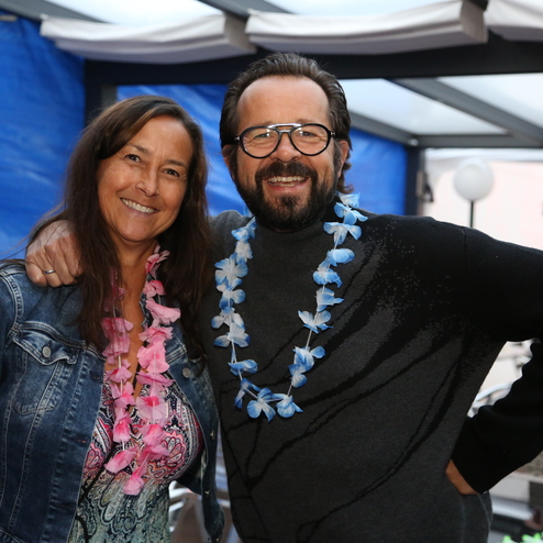 Zwei Personen mit farbigen Blumenketten l&auml;cheln in einem festlich geschm&uuml;ckten Raum.