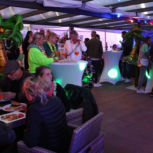 Viele G&auml;ste in festlicher Kleidung sitzen an Tischen und stehen an einer Bar in einem bunten, beleuchteten Veranstaltungsraum.