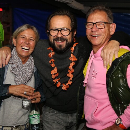 Drei Menschen lachen zusammen, mit einem Blumenkranz und bunten Shirts in einer festlichen Umgebung.