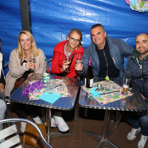 Gruppe von f&uuml;nf Personen mit Getr&auml;nken an einem Tisch, unter einer blauen Plane. Feierliche Atmosph&auml;re im Freien.