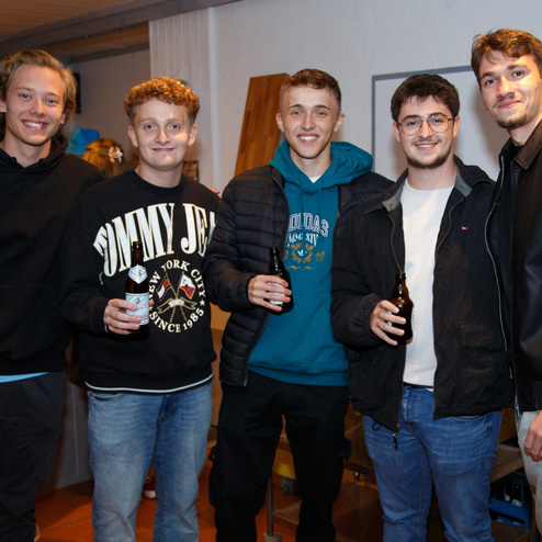F&uuml;nf junge M&auml;nner stehen l&auml;chelnd in einem Raum und halten jeweils ein Getr&auml;nk in der Hand.