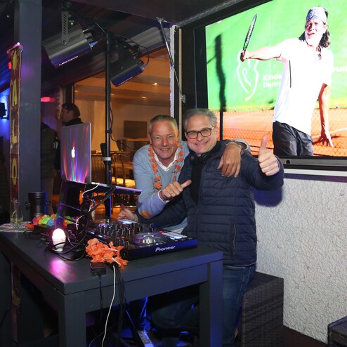 Zwei M&auml;nner stehen an einem DJ-Pult, w&auml;hrend im Hintergrund ein Tennis-Event auf einem Bildschirm gezeigt wird.