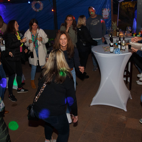 Menschen tanzen und feiern in einem festlich beleuchteten Raum mit einem Stehtisch und Getr&auml;nken.