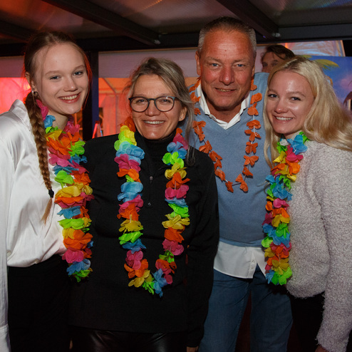 Vier Personen stehen zusammen und tragen bunte Blumenketten auf einer festlich geschm&uuml;ckten Party.