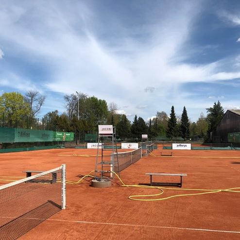 Tennisplatz aus rotem Sand mit Netzen, Bänken und Werbebannern, umgeben von Bäumen und klarem Himmel.