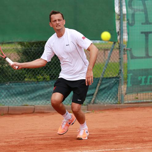 Ein Tennisspieler schlägt einen Ball mit einem Schläger auf einem roten Sandplatz. Hintergrund mit Netzen und grünen Bannern.