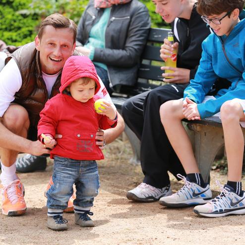Ein Mann hilft einem kleinen Jungen in roter Jacke, während weitere Kinder auf einer Bank sitzen und zusehen.