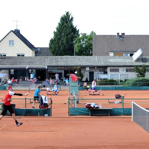 Tennisplatz mit Spielern in Aktion, Zuschauer im Hintergrund und Gebäude mit Terrasse. Rote Sandoberfläche, Netz sichtbar.