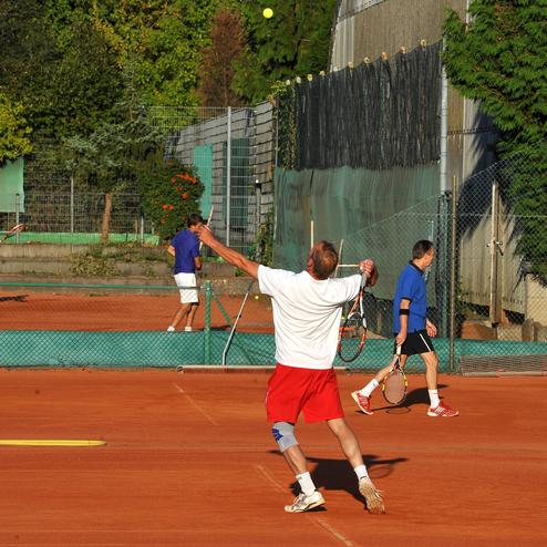 Vier Männer spielen Tennis auf einem Sandplatz, während ein Spieler den Aufschlag vorbereitet. Grünes Umfeld im Hintergrund.