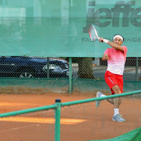 Tennisspieler schlägt den Ball mit einem Schläger auf einem orangefarbenen Sandplatz, im Hintergrund sind Bäume und Autos.