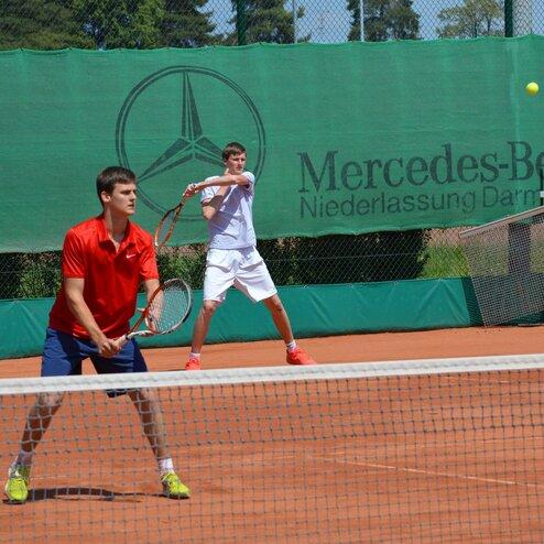 Zwei Spieler auf einem Tennisplatz: einer bereit zum Schlag, der andere in Spielhaltung, mit einem Netz im Vordergrund.