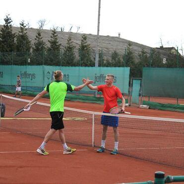 Zwei Tennis-Spieler schütteln sich nach einem Spiel die Hände, auf einem Sandplatz mit grünen Netzen im Hintergrund.