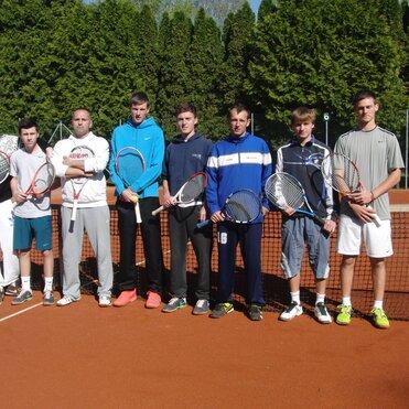 Gruppe von acht jungen Männern mit Tennisschlägern auf einem Sandplatz, hinter einem Netz und grünen Bäumen im Hintergrund.