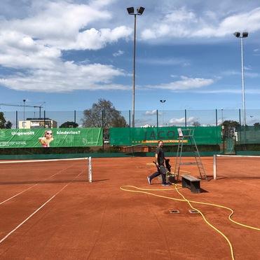 Tennisspielplatz mit einem Spieler, einem Schläger, einer Bank und Werbebanner im Hintergrund unter blauem Himmel.