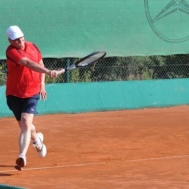 Mann in rotem Shirt schlägt einen Tennisball auf einem roten Sandplatz mit grüner Umrandung.