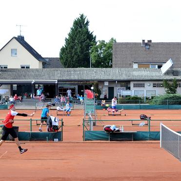 Tennisplatz mit Spielern in Aktion, Zuschauer im Hintergrund und Gebäude mit Terrasse. Rote Sandoberfläche, Netz sichtbar.