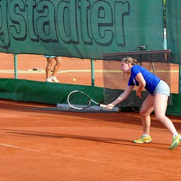 Tennisspielerin in blauer T-Shirt und Shorts bereit zum Schlag auf einem Tennisplatz mit rotem Sand.