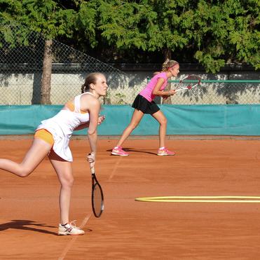 Zwei Tennis-Spielerinnen auf einem Sandplatz, eine schlagt den Ball, die andere bereitet sich vor.