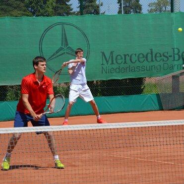 Zwei Spieler auf einem Tennisplatz: einer bereit zum Schlag, der andere in Spielhaltung, mit einem Netz im Vordergrund.