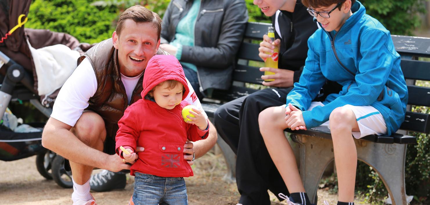 Ein Mann hilft einem kleinen Jungen in roter Jacke, w&auml;hrend weitere Kinder auf einer Bank sitzen und zusehen.