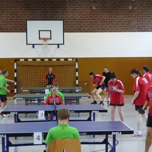 Tischtennistraining in einer Halle mit Spielern in roten und grünen Shirts an mehreren Tischen.