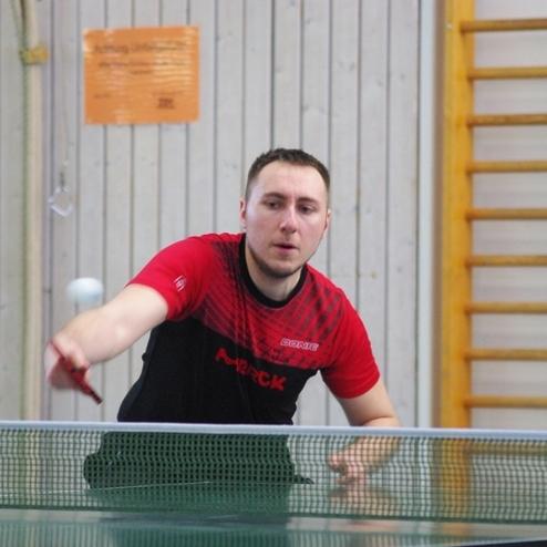 Tischtennisspieler in rotem Shirt schlägt einen Ball über das Netz auf einer Tischtennisplatte in einer Sporthalle.