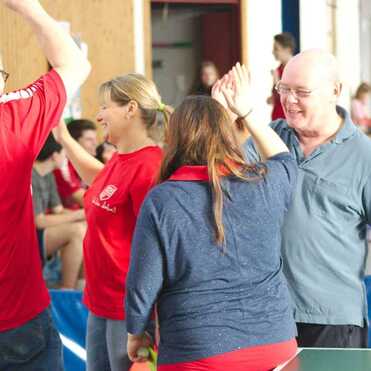 Vier Personen haben sich in einer Sporthalle um einen Tisch versammelt und freuen sich, während sie sich abklatschen.
